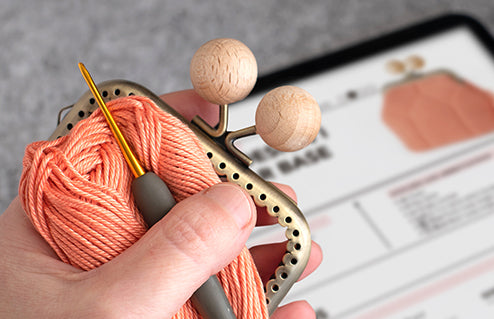 una mano sujeta gancho de crochet, hilo y boquilla para monedero y al fondo una tablet con el patron de crochet para hacer un monedero